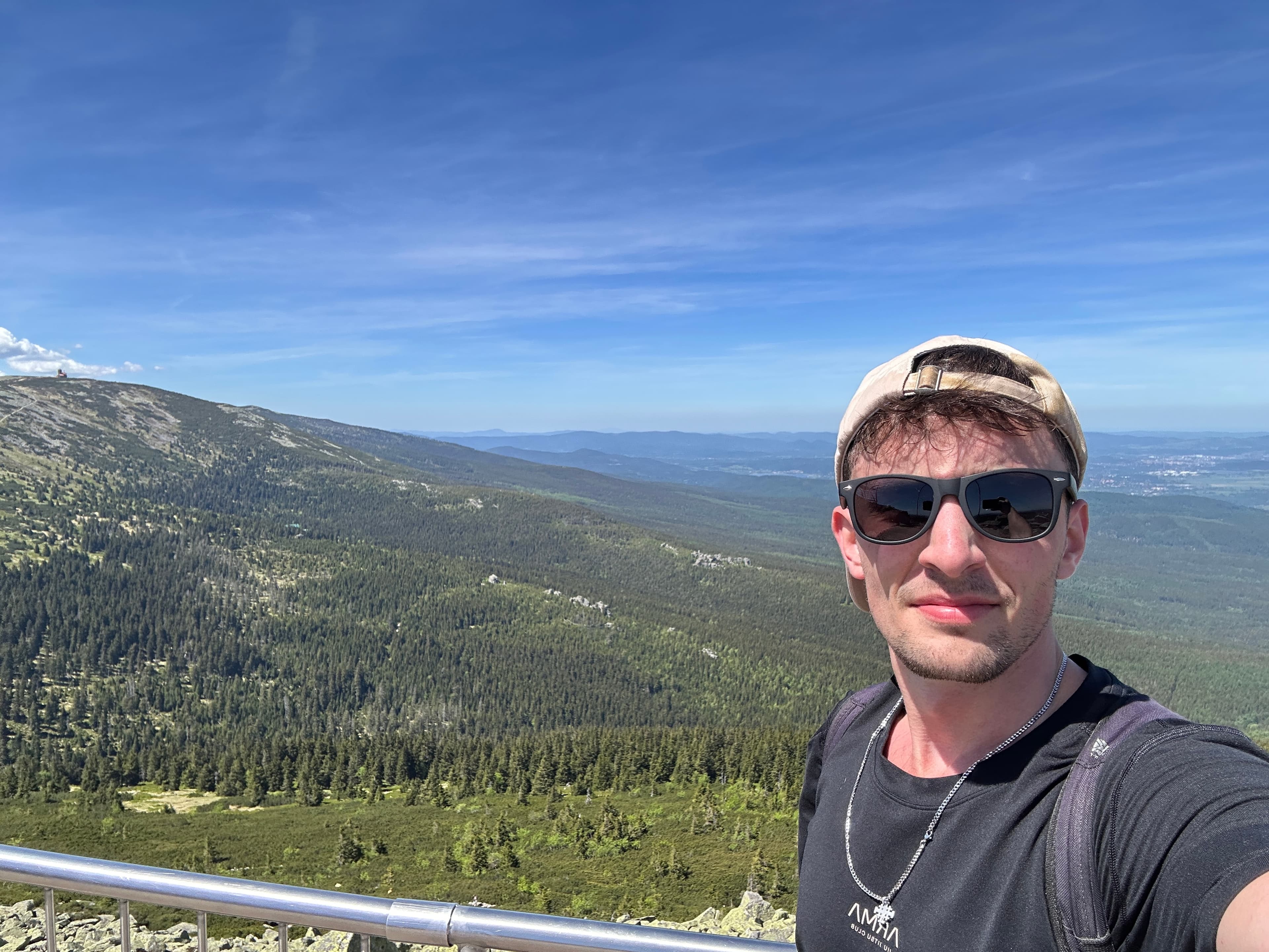 Nikodem taking a selfie on a mountain hike, in sunglasses and a cap, with forested ridges behind him
